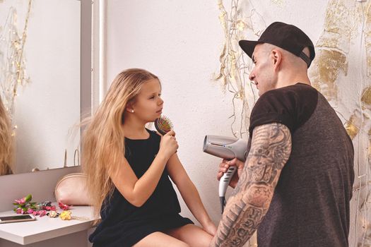 Cute little daughter and her tattoed dad are playing together near a mirror. Handsome dad is drying his daughter's hair while she is singing in the hairbrush. Family holiday and togetherness.