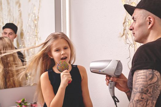 Cute little daughter and her tattoed dad are playing together near a mirror. Young dad is drying his daughter's hair while she is singing in the hairbrush and looking at the mirror. Family holiday and togetherness.