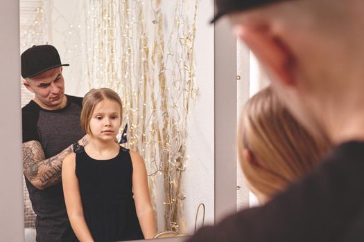 Cute little daughter and her tattoed dad are playing together near a mirror. Young dad is doing his daughter's hair. Family holiday and togetherness.