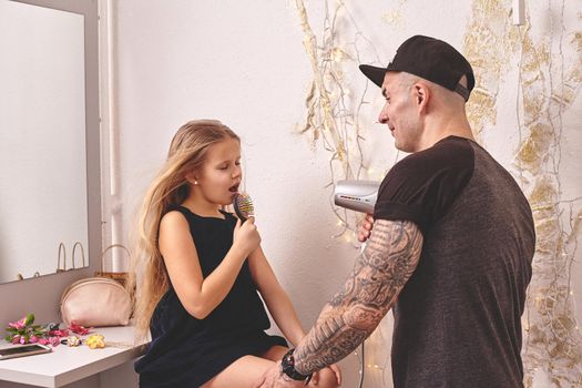 Cute little daughter and her tattoed dad are playing together near a mirror. Dad is drying his daughter's hair while she is singing in the hairbrush. Family holiday and togetherness.