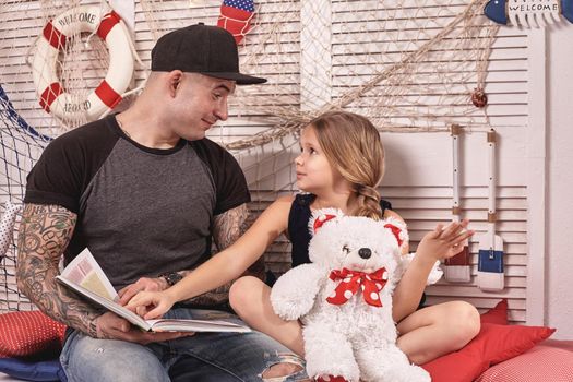 Handsome tattoed man in a cap is spending time with his little cute daughter. They are sitting on a white bench with some red pillows, in a room decorated in a marine style. She is holding a toy bear, pointing on something in a book and looking at her daddy. Reading fairytales while daughter is sitting nearby. Happy family.