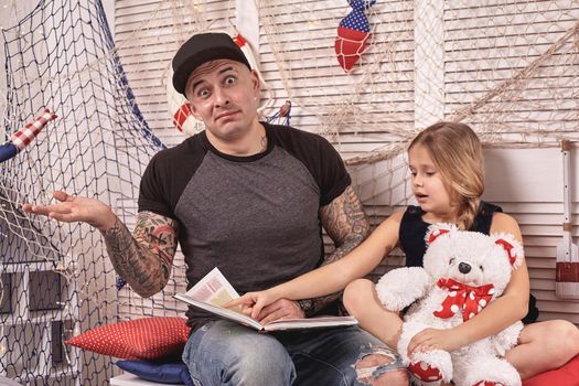 Handsome tattoed man in a cap is spending time with his little cute daughter. They are sitting on a white bench with some red pillows, in a room decorated in a marine style. She is holding a toy bear and pointing on something in a book. Reading fairytales and looking wondered while daughter is sitting nearby. Happy family.