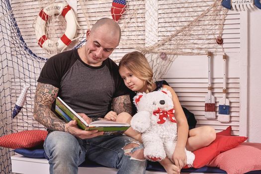 Bald tattoed man in a cap is spending time with his little cute daughter. They are sitting on a white bench with some red pillows, in a room decorated in a marine style. She is holding a toy bear. Reading fairytales while daughter is sitting nearby. Happy family.