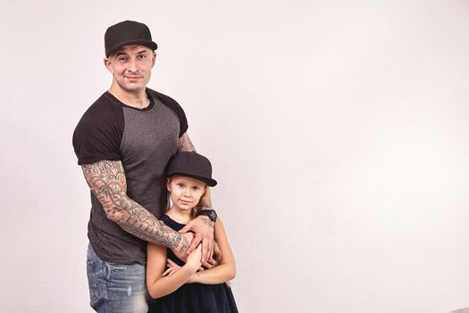 Cute little girl and her handsome tattoed father are looking at the camera and hugging, on a gray background. Each of them have a cap on their heads.