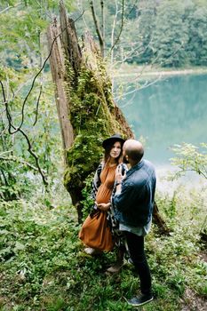 Man and a pregnant woman stand near a tree overgrown with moss by the lake. High quality photo