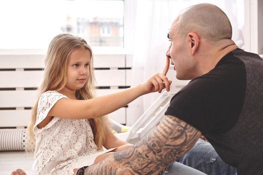 White modern kid's room whith a wooden furniture. Adorable daughter wearing a white dress is touching daddy's nose and looking with tenderness at him. Young daddy with tattoos is hugging her. Friendly family spending their free time together sitting on a pillows.