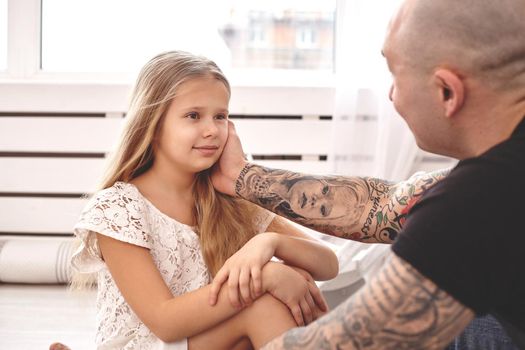 White modern kid's room whith a wooden furniture. Adorable daughter wearing a white dress is looking with tenderness at her loving father. Daddy with tattoos is stroking her cheek. Friendly family spending their free time together sitting on a pillows.