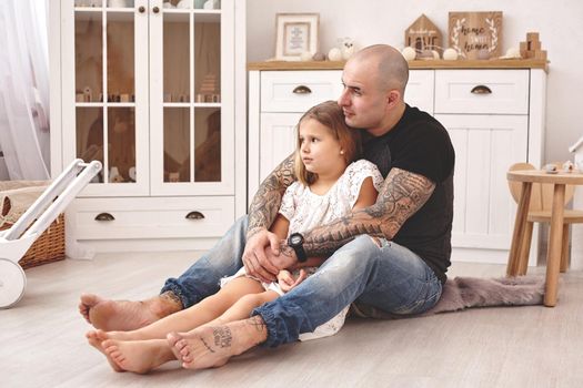 White modern kid's room whith a wooden furniture. Adorable daughter with a long blond hair wearing a white dress. Daddy with tattoos is hugging her and they are looking away. Friendly family spending their free time together sitting on a pillows.
