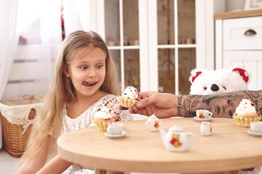 White modern kid's room whith a wooden furniture. Adorable daughter whith a long blond hair wearing a white dress. They are drinking tea from a toy dishes in a modern kid's room whith a wooden furniture. Daddy with tattoos is treating her with a cupcacke. Friendly family spending their free time together sitting on a pillows.