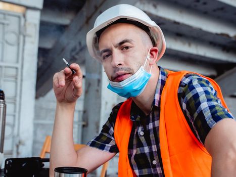 A concerned male civil engineer in a protective medical mask sits at a table at a construction site. Smoke break and reflection