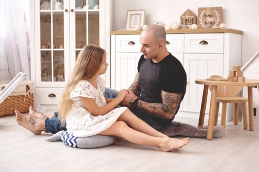 White modern kid's room whith a wooden furniture. Adorable daughter wearing a white dress is looking with tenderness at her loving father. Daddy with tattoos is holding her hands. Friendly family spending their free time together sitting on a pillows.