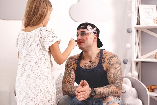 Funny time Tattoed father in a cap and his child are playing at home. Pretty girl in a white dress is putting a lipstick at her dady's lips, in her bedroom. Family holiday and togetherness.