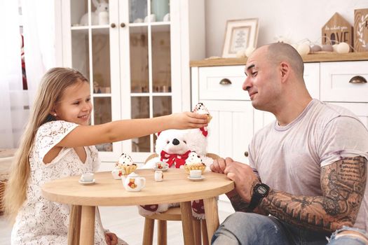 White modern kid's room whith a wooden furniture. Adorable daughter whith a long blond hair wearing a white dress is treating her daddy with a cupcacke. They are drinking tea from a toy dishes in a modern kid's room whith a wooden furniture. Daddy with tattoos is looking happy. Friendly family spending their free time together sitting on a pillows.