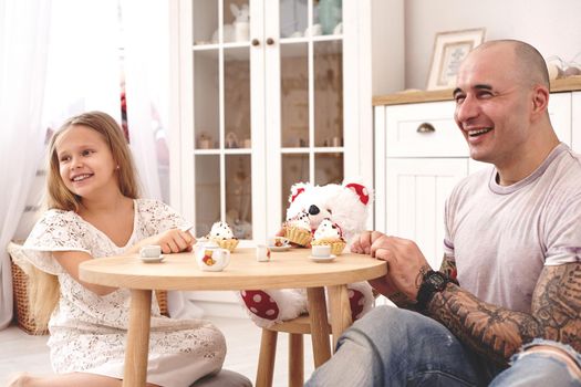 White modern kid's room whith a wooden furniture. Adorable daughter whith a long blond hair wearing a white dress. They are drinking tea with cupcackes from a toy dishes in a modern kid's room whith a wooden furniture. Daddy with tattoos is laughing with his daughter. Friendly family spending their free time together sitting on a pillows.