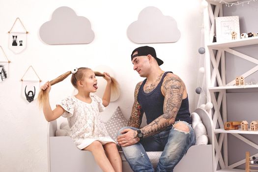 Funny time Tattoed father in a cap and his child are playing at home. Dad is doing his daughter's hair in her bedroom. They are looking at each other. Family holiday and togetherness.