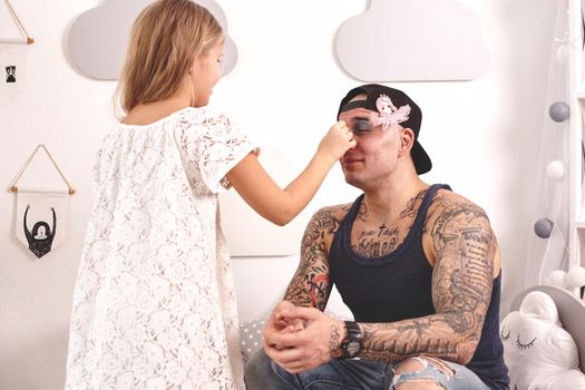 Funny time Tattoed father in a cap and his child are playing at home. Pretty girl in a white dress is doing makeup to her dad in her bedroom. Family holiday and togetherness.