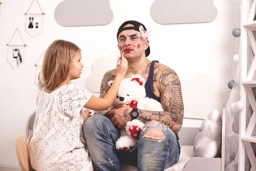 Funny time Tattoed father in a cap and his child are playing at home. Pretty girl in a white dress is putting a lipstick at her dady's lips while he is holding a toy bear, in the bedroom. Family holiday and togetherness.