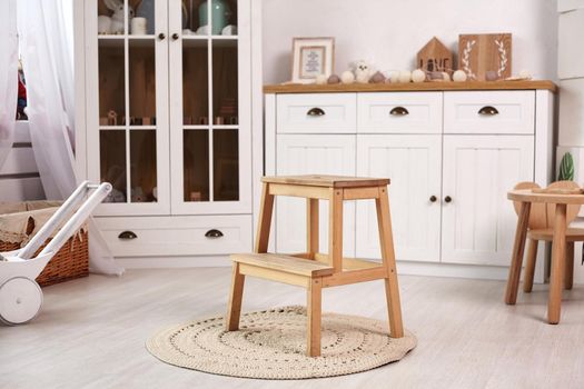 Elegant kid's room with a white wooden furniture and a window. Small cute heart shaped chair and table, little wooden ladder on a round rug in the middle of the room. White decorative wheelbarrow and a pad near the window.