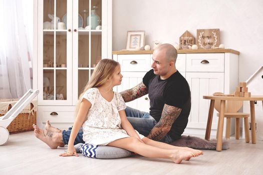 White modern kid's room whith a wooden furniture. Adorable daughter wearing a white dress is looking with tenderness at her loving father. Daddy with tattoos is hugging her. Friendly family spending their free time together sitting on a pillows.