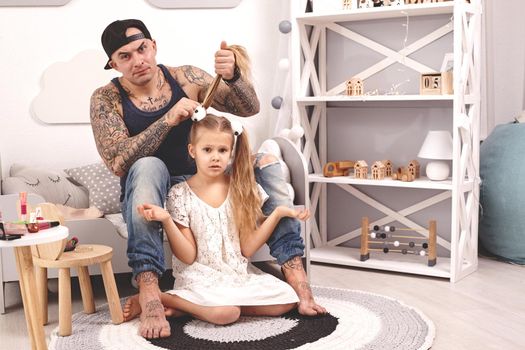 Funny time Tattoed father in a cap and his child are playing at home. Dad is doing his daughter's hair while she is sitting on a rug in her bedroom. Family holiday and togetherness.