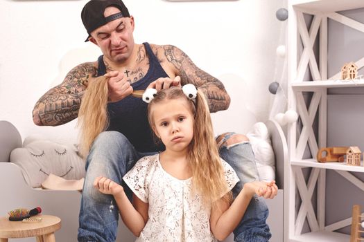 Funny time Tattoed father in a cap and his child are playing at home. Dad is doing his daughter's hair in her bedroom. Family holiday and togetherness.