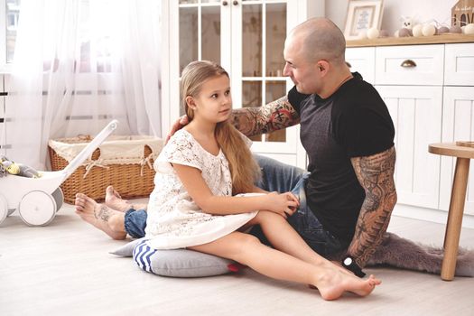 White modern kid's room whith a wooden furniture. Adorable daughter wearing a white dress is looking with tenderness at her father. Daddy with tattoos is hugging her. Friendly family spending their free time together sitting on a pillows.