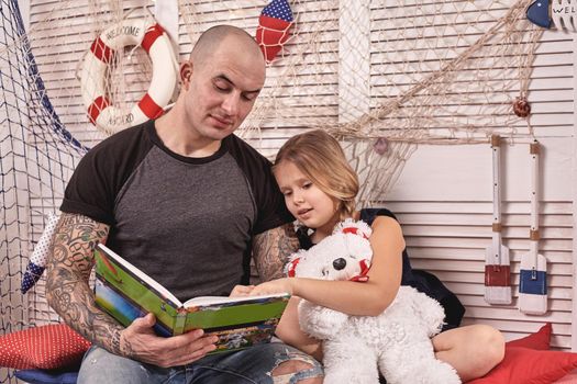 Athletic tattoed man in a cap is spending time with his little cute daughter. They are sitting on a white bench with some red pillows, in a room decorated in a marine style. She is holding a toy bear and looking at the book. Reading fairytales while daughter is sitting nearby. Happy family.