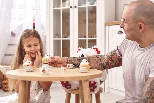 White modern kid's room whith a wooden furniture. Adorable daughter wearing a white dress with a long blond hair. They are drinking tea from a toy dishes in a modern kid's room whith a wooden furniture. Young daddy with tattoos is treating his daughter with a cupcacke. Friendly family spending their free time together sitting on a pillows.