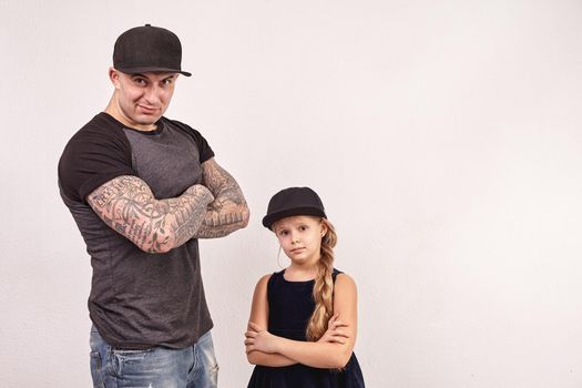 Cute little girl and her handsome tattoed father have crossed their hands and looking at the camera on a gray background. Each of them have a cap on their heads.