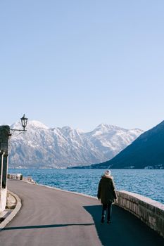 Girl in a jacket walks along the asphalt embankment against the background of the sea and mountains. High quality photo