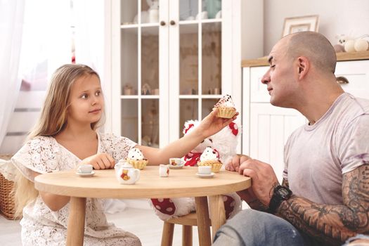 White modern kid's room whith a wooden furniture. Adorable daughter whith a long blond hair wearing a white dress is treating her daddy with a cupcacke. They are drinking tea from a toy dishes in a modern kid's room whith a wooden furniture. Daddy with tattoos is going to try a treatment. Friendly family spending their free time together sitting on a pillows.