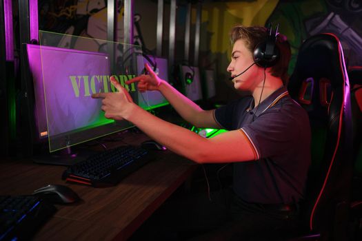 Photo of professional young caucasian gamer playing online on his PC in gaming club or cyber arena. he is upset and shows rude gestures to his virtual opponents. laser screen, projector, augmented reality, AR, VR