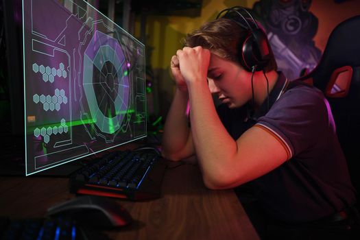 Photo of professional young caucasian gamer playing online on his PC in gaming club or cyber arena. He looks displeased and upset. Game plan went wrong. laser screen, projector, augmented reality, AR, VR