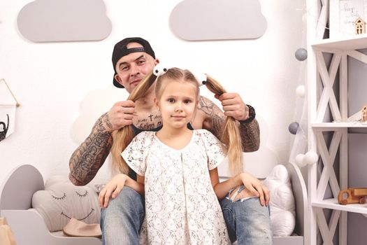 Funny time Tattoed father in a cap and his child are playing at home. Dad is doing his daughter's hair in her bedroom. They are looking at the camera and smiling. Family holiday and togetherness.