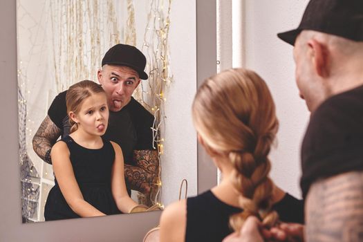 Cute little daughter and her tattoed dad are playing together near a mirror. Dad is doing a braid for his little princess. They are showing tongues to each other. Family holiday and togetherness.