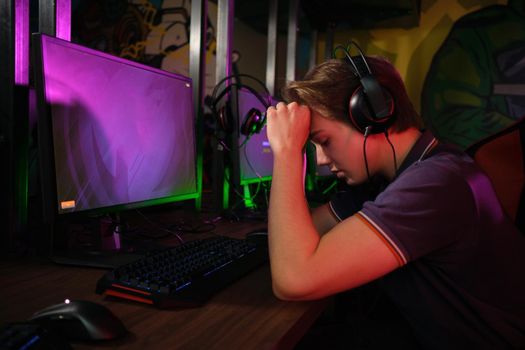 Photo of professional young caucasian gamer playing online on his PC in gaming club or cyber arena. He looks displeased and upset. Game plan went wrong