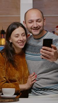 Cheerful couple using video call communication on smartphone talking to family on christmas eve day. Man and woman with remote conversation on online conference for holiday season