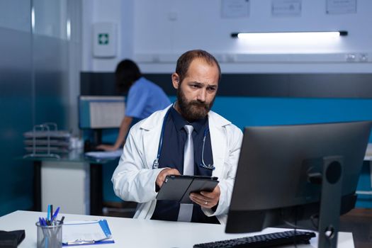 Specialist using technology for healthcare system and practice at office. Doctor holding digital tablet while looking at computer screen for analysis information, working late.