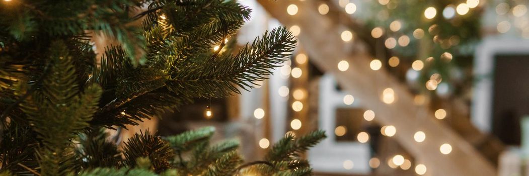 Home New Year's interior. A live Christmas tree and a wooden staircase in the kitchen. Cozy atmosphere of a home holiday, loft design of the room, daylight, green tree decorated with lights. Modern Scandinavian style in the interior. The eve of December 25th.