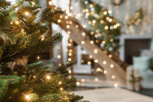 Home New Year's interior. A live Christmas tree and a wooden staircase in the kitchen. Cozy atmosphere of a home holiday, loft design of the room, daylight, green tree decorated with lights. Modern Scandinavian style in the interior. The eve of December 25th.