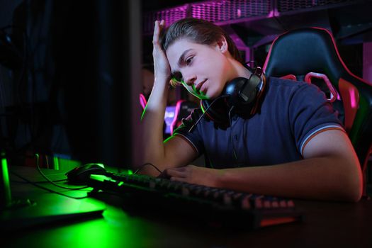 Photo of professional young caucasian gamer playing online on his PC in gaming club or cyber arena. He looks displeased and upset. Game plan went wrong