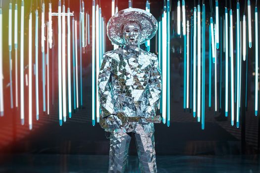 A male artist in a mirrored suit and hat with an electric guitar on a dark background, space for text