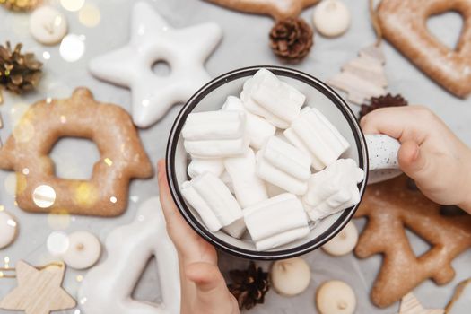 Children's hands hold a cup of cocoa with marshmallows. Christmas gingerbread on the table, bokeh lights in the foreground. The concept of desserts and drinks during the Christmas holidays.