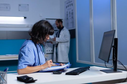 Woman nurse doing overtime work with computer and checkup files. Medical assistant using information papers and monitor for healthcare system. Adult with expertise working late.