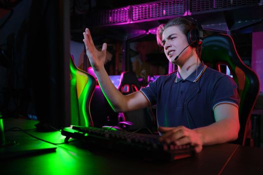 Photo of professional young caucasian gamer playing online on his PC in gaming club or cyber arena. He looks displeased and upset. Game plan went wrong