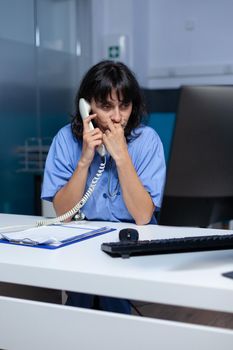 Medical assistant talking on landline phone to patient for appointment, working late. Nurse using telephone for remote communication and healthcare system while sitting at desk.