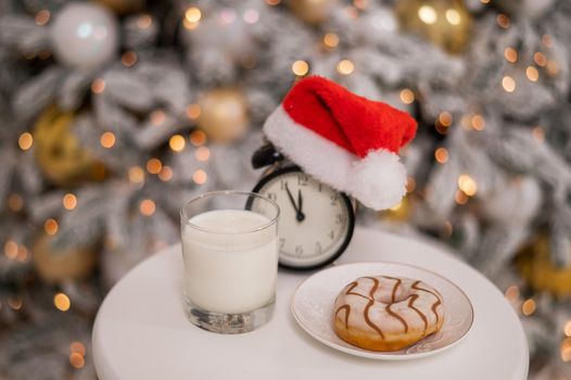 Alarm clock in a santa claus hat with a glass of milk and a donut on the background of a christmas tree