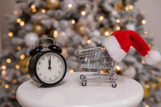 Christmas gifts shopping time. A shopping trolley with a santa hat and an alarm clock by the Christmas tree