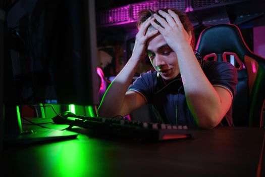 Photo of professional young caucasian gamer playing online on his PC in gaming club or cyber arena. He looks displeased and upset. Game plan went wrong