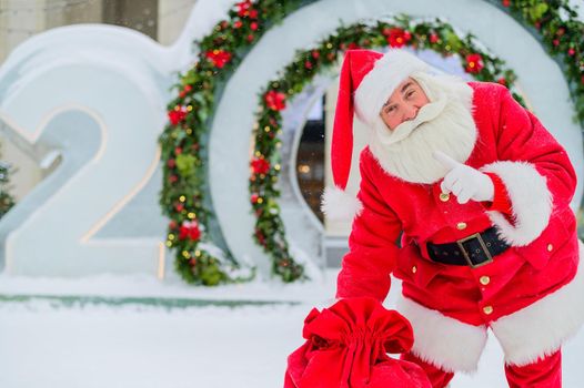 Santa Claus posing with a bag of gifts on the background of Christmas decorations outdoors.